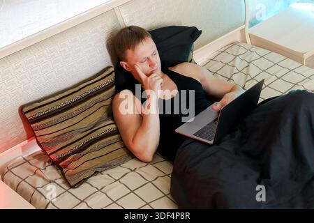 Ein besinnlicher Mann liegt auf einem Bett mit bunten Kissen, Hand am Kinn und tippt auf einem eleganten Laptop unter warmen Umgebungslichtern in einem stilvollen Zimmer. Stockfoto