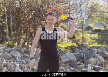 Starker Erwachsener in Sportbekleidung hebt den Arm mit lebhaftem Schläger auf der Schulter, Hand an Ball, in natürlicher felsiger Landschaft. Stockfoto