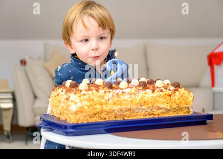 Ein glückliches Kleinkind steht drinnen vor einer Geburtstagskuche, während es während einer Familienfeier eine beleuchtete Kerze ausbläst. Stockfoto