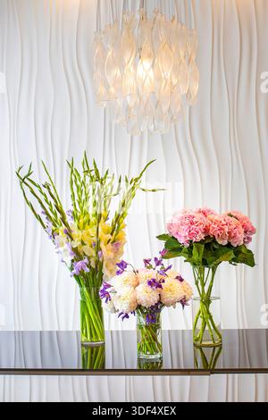 Drei Blumenarrangements in Glasvasen auf einem schwarzen Glastisch vor einer weißen welligen Wand, beleuchtet von einer Kristall-Pendelleuchte. Stockfoto