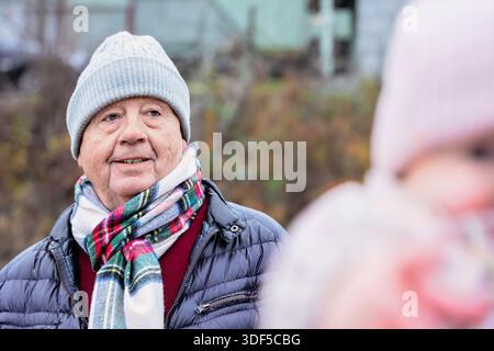 Seniorenmann in warmer Kleidung, lächelnd im Freien, umgeben von der Natur, fängt eine fröhliche Atmosphäre bei kaltem Wetter ein Stockfoto