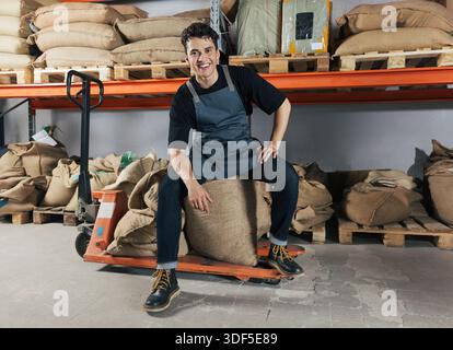 Selbstbewusster und fröhlicher Lagerarbeiter, der auf der Tasche sitzt. Junger lächelnder männlicher Arbeiter auf einer Schürze posiert in einem Lagerhaus in Regalen Stockfoto