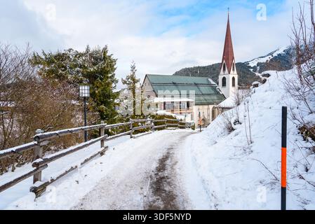 Umzäunter schneebedeckter Wanderweg in einem Bergdorf in Tirol an einem Wintertag. Eine traditionelle Kirche mit einem hohen Glockenturm ist im Hintergrund zu sehen. Stockfoto