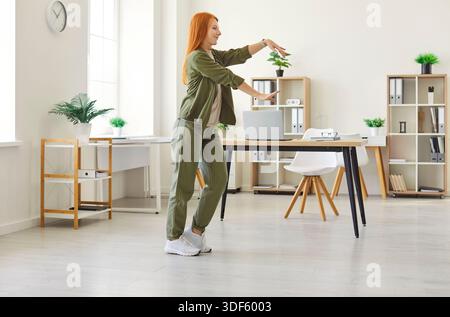 Fröhliche rothaarige Frau in Khaki-Outfit tanzt am Arbeitsplatz, genießt Pause, Balance zwischen Arbeit und Sport. Stockfoto
