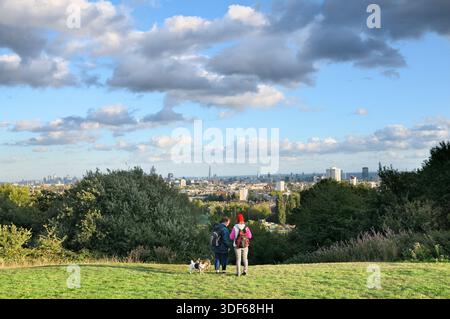Zwei Frauen mit Hunden halten für einen Moment an, um die Aussicht auf Parliament Hill mit Blick auf die Skyline von London, Hampstead Heath, London, England, Großbritannien zu genießen. Stockfoto