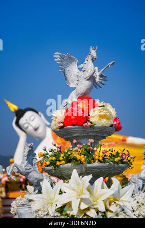 Weiße Pegasus-Statue auf einem floralen Sockel mit einem liegenden Buddha im Hintergrund im Wat Phra That Doi Kham Tempel auf der Bergseite von Chiang Ma Stockfoto