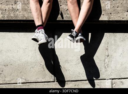 Zwei Personen, die an einer Wand sitzen und die Beine schwingen. Bild nur Beine. Stockfoto