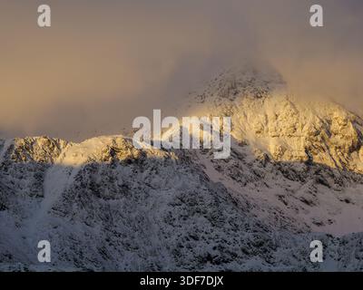Ein schneebedeckter Berggrat, der von Wolken verdeckt ist und einen Teil davon von Sonnenlicht beleuchtet, Yr Eryri National Park, Wales, Januar 2026 Stockfoto