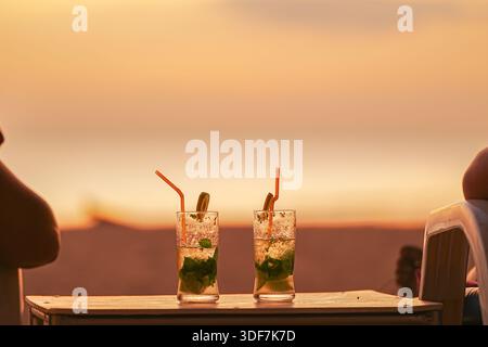 Zwei Mojito-Cocktails, die bei Sonnenuntergang in Gläsern serviert werden. Ein Paar Urlaub in Resorts und Unterhaltung, eine Flitterwochenreise. Zwei Leute beobachten die Stockfoto