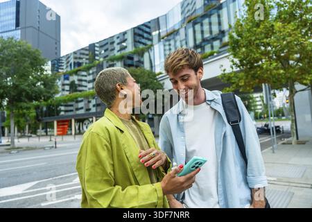 Zwei verschiedene Kollegen, die sich in lockeren Gesprächen unterhalten und auf einer städtischen Straße einen Smartphone-Bildschirm teilen, mit moderner Stadtarchitektur und Grün Stockfoto