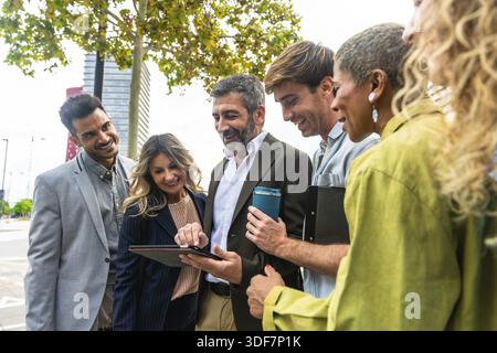 Verschiedene Gruppen lächelnder Geschäftsleute betrachten gemeinsam einen Tablet-Bildschirm, diskutieren Ideen und planen Arbeit, während sie außerhalb eines Bürogebäudes stehen Stockfoto