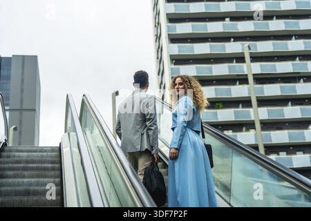 Zwei verschiedene Geschäftskollegen, die eine Rolltreppe hinaufsteigen, blicken in Richtung eines modernen Stadtgebäudes, das Karrierewachstum, Zusammenarbeit und städtische PR symbolisiert Stockfoto