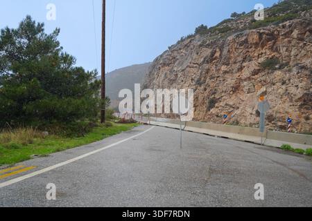 Geschlossene Ausfahrt zum North Cretan Highway (BOAK) in Milopotamos. Betonbarrieren und Verkehrsschilder blockieren die Straße neben einer steilen felsigen Klippe Stockfoto