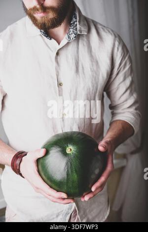 Bärtiger Mann im Leinenhemd hält in seinen Händen ganz grüne Wassermelone Stockfoto