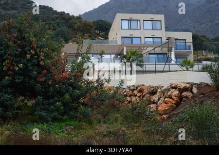 Luxuriöse, minimalistische Villa mit großen Glasfenstern und Terrasse. Moderne High-End-Architektur an einem steilen kretischen Hang mit tropischen Pflanzen und Felsen Stockfoto