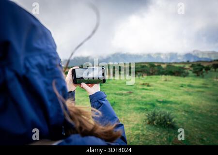Rückansicht einer Frau, die mit einem Smartphone eine grüne Landschaft filmt, Reise- und Content-Creation-Konzept mit Kopierraum. Stockfoto