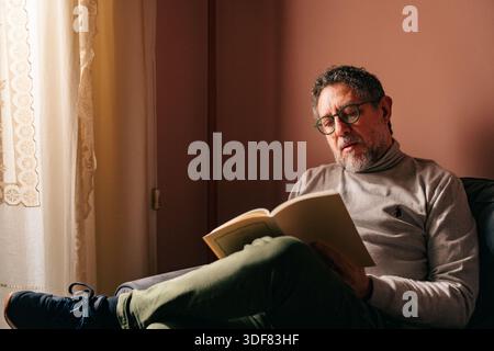 Mann mittleren Alters, der zu Hause ein Buch auf dem Sofa liest, tief konzentriert und vom Lesen vertieft. Ruhige Innenaufnahme mit warmem Licht, die Konzentration ausdrückt, Stockfoto