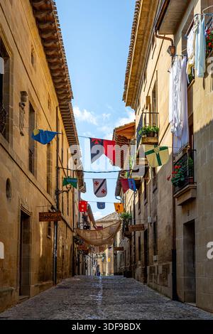 Sonnendurchflutete Kopfsteinpflasterstraße in einer historischen europäischen Stadt flattern bunte Wimpelfahnen zwischen Steinhäusern, Briones, La Rioja, Spanien Stockfoto