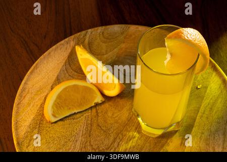 Ein Glas frisch gepressten Orangensaft mit einem Glasstroh und Orangenscheiben und Hälften, die daneben auf einem Holztablett liegen. Stockfoto
