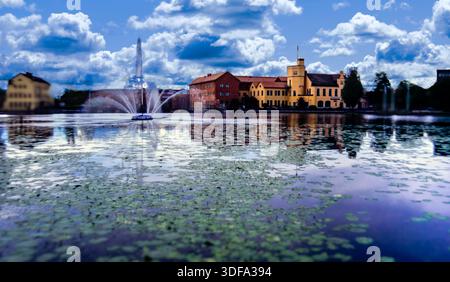 Blick bei Dämmerung auf ein mit Seerosen bedecktes Rive mit einem Brunnen und gelben Gebäuden im Hintergrund in Eskilstuna, Schweden. Stockfoto
