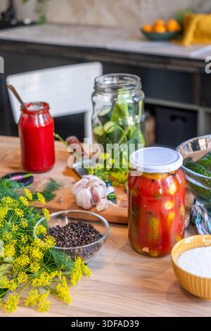 Gläser mit eingelegten Gurken und Knoblauch werden auf einem Holztisch ausgestellt, umgeben von frischem Dill und Gewürzen, die den Prozess der Gemüsepresse hervorheben Stockfoto