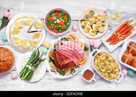 Klassisches Osterschinken-Abendessen. Tischszene mit Blick von oben auf weißem Holzhintergrund. Schinken, Muschelkartoffeln, Beilagen mit Gemüse, Eier, Brot und lemo Stockfoto