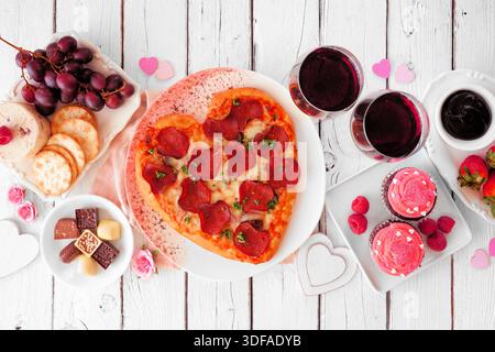 Hausgemachtes Valentinstag-Abendessen. Tischszene mit Draufsicht auf weißem Holzhintergrund. Herzförmige Pizza, Pasta, Wein, Käseplatte und Desserts. Stockfoto