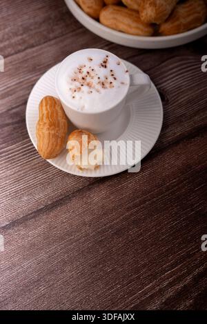 Blick von oben auf einen schäumenden Cappuccino in einer weißen Tasse und Untertasse mit Kakaostreuseln, begleitet von cremegefüllten Eclairs auf einem rustikalen braunen Holzkorn Stockfoto