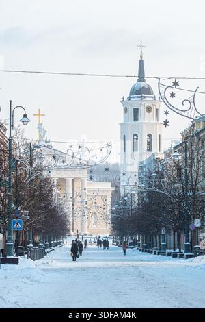 Schneebedeckte Fußgängerzone in Vilnius, Litauen, mit Menschen, die auf der Hauptstraße spazieren gehen, dekoriert mit festlichen Lichtern in einer winterlichen Stadtatmosphäre Stockfoto