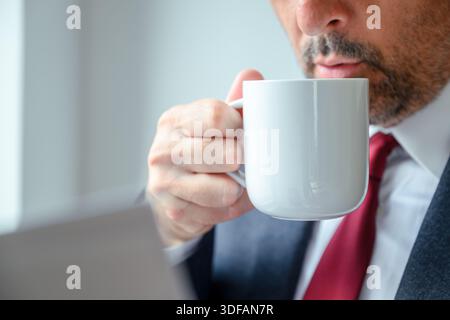 Professioneller Mann in Anzug, der Kaffee aus einer Tasse am Fenster trinkt, konzentrieren Sie sich auf eine Tasse in eleganter Büroumgebung. Selektiver Fokus. Stockfoto