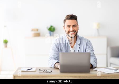 Erfolgreicher Freiberufler. Glücklicher junger Mann, der mit einem Laptop am Arbeitsplatz sitzt und vor der Kamera lächelt und Platz kopiert Stockfoto