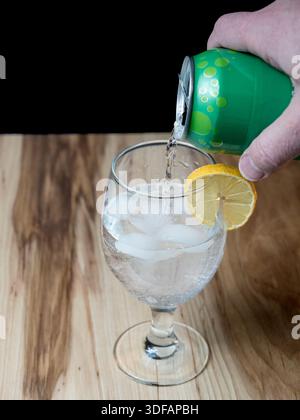 Ein Hochwinkelfoto erfrischendes Limonade aus einer grünen Dose in ein mit Eis und einer Zitronenscheibe gefülltes Glas auf einer Holzoberfläche. Stockfoto