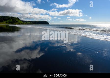 Wellen schlagen sanft gegen einen ruhigen schwarzen Sandstrand unter einem hellblauen Himmel. Flauschige Wolken reflektieren das Wasser und schaffen eine friedliche Atmosphäre Stockfoto