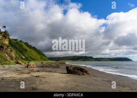 Wellen schlagen sanft gegen den dunklen Sandstrand, der von hohen Klippen und lebhaften grünen Hügeln unter einem dramatischen Himmel eingerahmt wird. Eine ruhige Atmosphäre lädt zum Verweilen ein Stockfoto