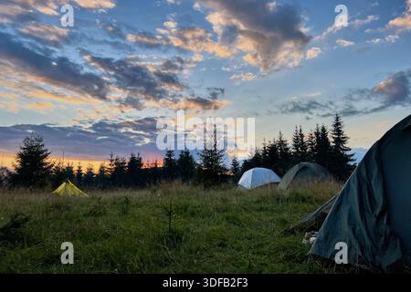 Wenige Touristenzelte in einem grasbewachsenen Feld am Abend, umgeben von Bäumen und Hügeln unter atemberaubendem Sonnenuntergangshimmel. Farbenfrohe Wolken verleihen der Szene eine schöne Note mit leuchtenden Orange- und Violetttönen am Horizont. Stockfoto