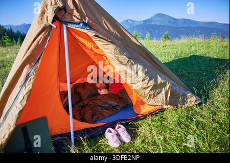 Das Kind schläft friedlich im orangefarbenen und braunen Zelt, kuschelig im Schlafsack auf blauer Matte. Sonnenlicht filtert ein und erhellt ihr ruhiges Gesicht. Karpaten, Ukraine. Stockfoto