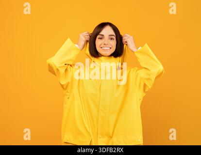 Glückliche Frau in gelber wasserdichter Regenjacke, die Kapuze aufsetzt und isoliert auf gelbem Hintergrund steht Stockfoto