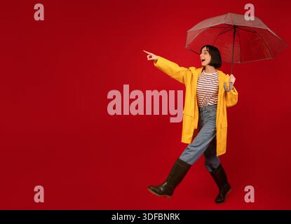 Verkaufskonzept Herbst. Überraschte Dame in wasserdichtem Regenmantel, die Regenschirm hält und leeren Raum auf rotem Hintergrund zeigt Stockfoto