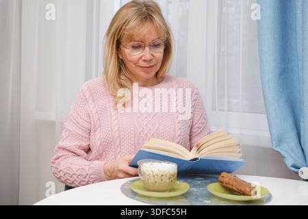Reife Frau, die zu Hause ein Buch liest, während sie Tee am Esstisch trinkt, gemütliche Abendeinrichtung mit Laptop in der Nähe Stockfoto