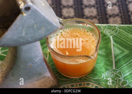 Frisch gepresster Orangensaft, der aus der manuellen Metallpresse in eine Glasschale auf einem grünen Tisch mit grünem Muster gefüllt wird und erfrischend gesundes Zitrusgetränk voller Zitrusfrüchte bietet Stockfoto