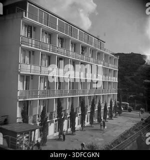 Krim, Ukraine, UdSSR, 1960er Jahre Ein stimmungsvolles Schwarzweiß-Archivfoto, das ein modernes mehrstöckiges Sanatorium oder Hotelgebäude erfasst. Die Fassade hat durchgehende Balkonreihen und große Fenster, typisch für die frühe sowjetische Moderne. Eine Reihe junger Zypressen ziert den Eingangsbereich. Auf der rechten Seite parkt ein Oldtimer-Bus vor einem Hang. Die Leute schlendern durch den sonnigen Innenhof. Auf dem Dach befindet sich ein verglastes Solarium. Diese Ansicht veranschaulicht die Entwicklung der Resortinfrastruktur an der Südküste während einer friedlichen Zeit. Stockfoto