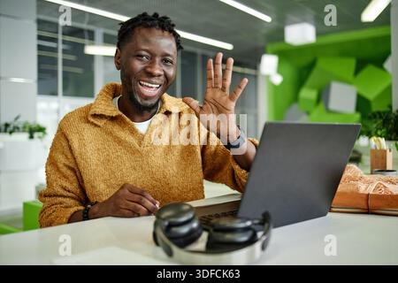 Porträt eines jungen Erwachsenen Schwarzen Mannes, der mit Laptop am Schreibtisch lächelt und winkt Stockfoto