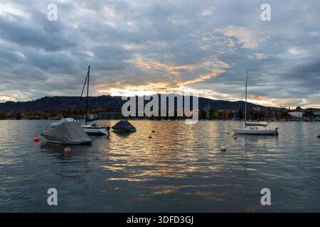 Blick auf den Sonnenuntergang im Herbst über den berühmten Züricher See mit Blick auf die herbstfarbenen Kiefern auf den Schweizer Alpen mit einigen Sonnenstrahlen und den Booten und Stockfoto