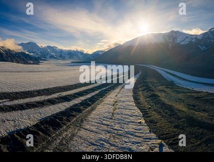 Aus der Vogelperspektive auf den dunklen, gewundenen Pfad, der durch die unberührte, schneebedeckte Landschaft unter den hoch aufragenden, schneebedeckten Bergen unter einem strahlenden Licht führt Stockfoto