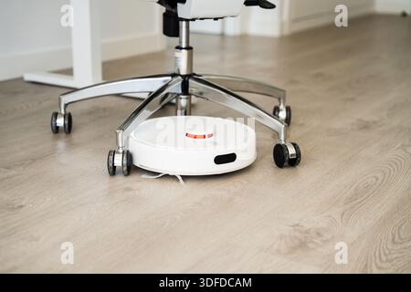 Intelligenter Roboter-Staubsauger, der unter Bürostuhl auf Holzboden arbeitet, automatisches Reinigungsgerät für moderne Heim- und Arbeitsbereiche Stockfoto