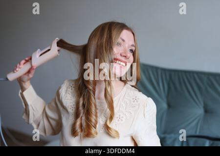 Lifestyle-Schönheitsbild einer lächelnden Frau mit Haarstyling-Werkzeug in minimalem gemütlichem Innenraum Stockfoto