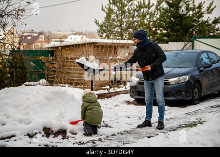 Mann schaufelt Schnee im Garten, Kind hilft mit einer Spielzeugschaufel. Stockfoto