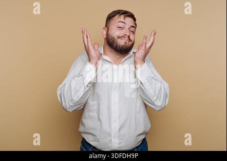 Ein Mann steht in einem Studio mit weißem Hemd und blauer Jeans. Er lächelt und zuckt mit einem entspannten Ausdruck mit den Schultern. Der Hintergrund ist einfach Stockfoto