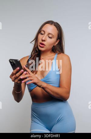 Eine Frau steht drinnen in Workout-Kleidung, während sie auf ihr Smartphone schaut. Sie scheint auf dem Bildschirm konzentriert zu sein, wenn sie das Gerät berührt. Die Szene-Show Stockfoto