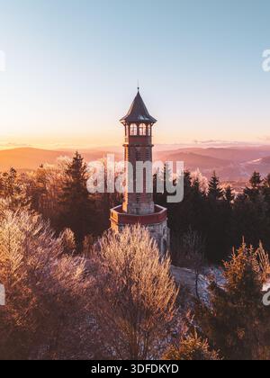 Der Stepanka-Aussichtsturm steht im Winter im Isergebirge hoch. Frost bedeckt die Bäume, während die Sonne untergeht, und wirft warmes Licht auf die Landschaft. Eine friedliche Szene lädt zum Erkunden ein. Stockfoto